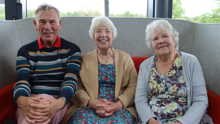 An elderly man and two women sitting on a grey sofa smiling