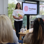 Young white woman with long dark hair in a treetops Hospice t-shirt giving a talk about the hospice to a local group