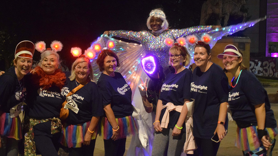 An LED stilt walker brightly lit behind a group of smiling women wearing Moonlight Walk t-shirts