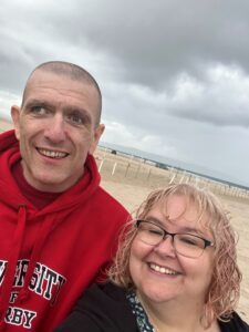 Man with red jumper and woman with glasses standing on a beach - Dom and Tracey