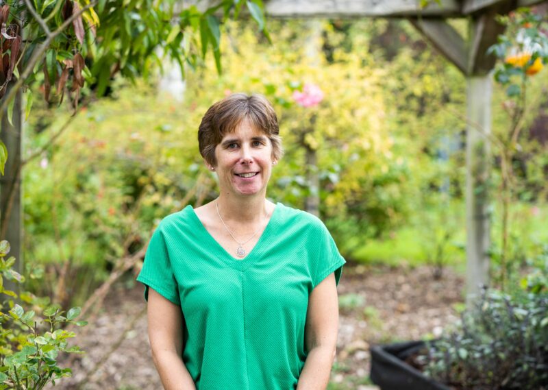 A person wearing a green shirt stands outdoors, surrounded by lush greenery and blooming plants, creating a serene garden atmosphere.